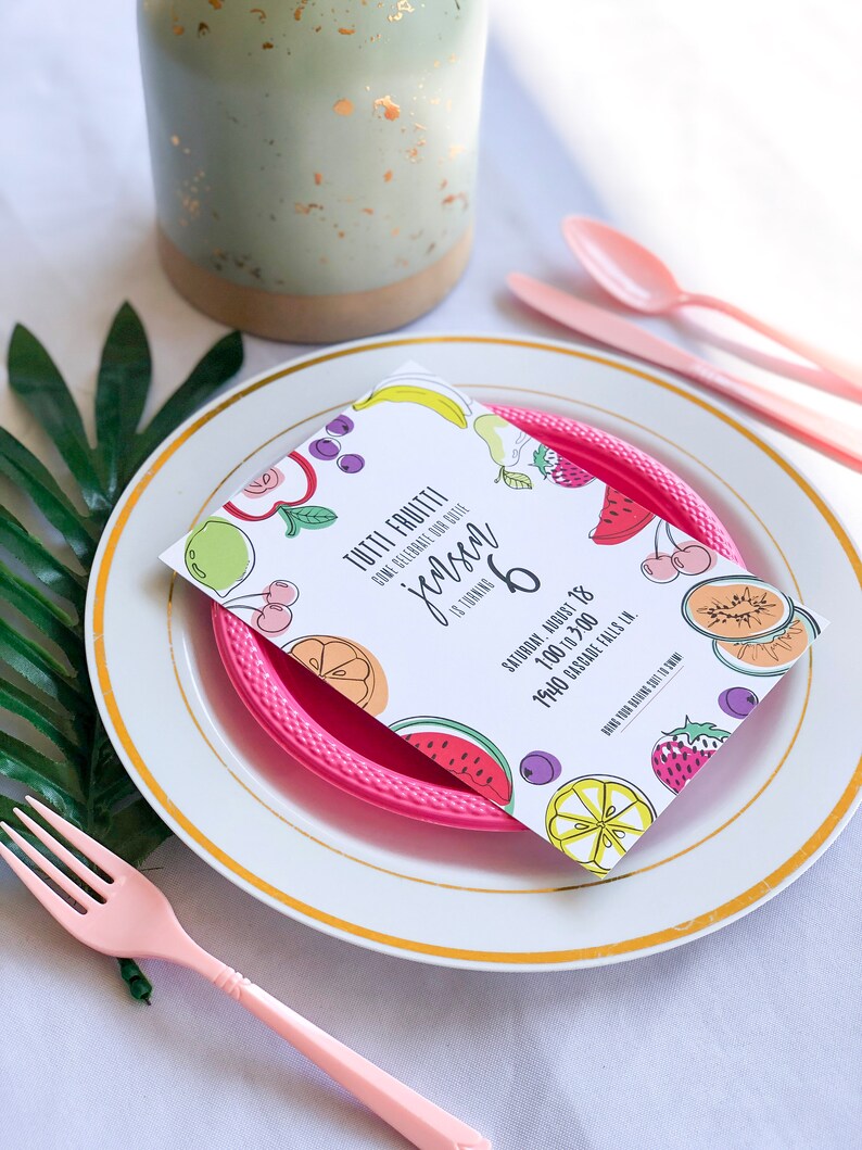 Puede incluir: Una tarjeta blanca con un dise&ntilde;o de frutas coloridas y el texto "Tutti Frutti" y "Jensen cumple 6 a&ntilde;os". La tarjeta est&aacute; sobre un plato de pl&aacute;stico rosa con un plato blanco debajo. El plato est&aacute; sobre un mantel blanco con una hoja verde en el fondo.