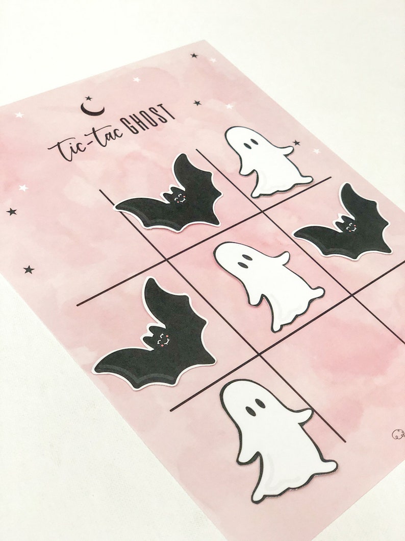 May include: A pink and white tic-tac-toe game board with a black bat and white ghost game pieces. The game board has the words "tic-tac ghost" written on it.