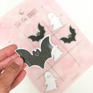 May include: A black bat sticker with a smiling face. The sticker is being held in a hand. The sticker is for a tic-tac-toe game with a pink background and white ghost and bat stickers.