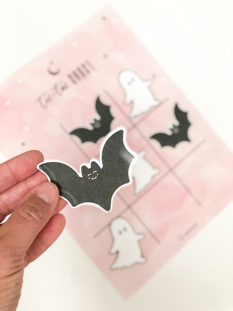 May include: A black bat sticker with a smiling face. The sticker is being held in a hand. The sticker is for a tic-tac-toe game with a pink background and white ghost and bat stickers.