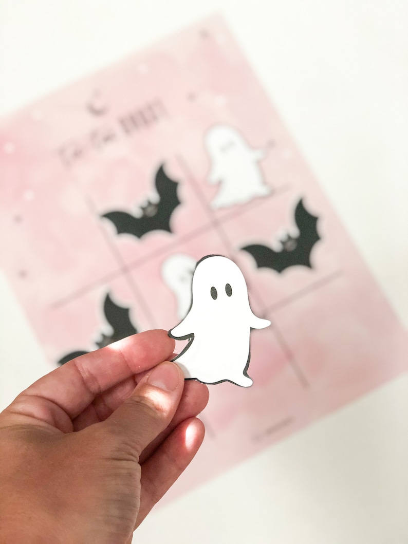 May include: A white ghost cutout with black eyes and a black outline. The ghost is being held by a hand in front of a pink and white background with black bats and ghosts.
