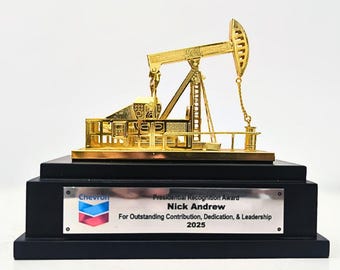 May include: A gold-colored oil pump jack award on a black base. The award has a plaque with the Chevron logo and the text "Presidential Recognition Award Nick Andrew For Outstanding Contribution, Dedication, & Leadership 2025."
