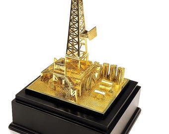 May include: A gold-colored model of an oil rig, featuring a tall derrick and detailed platform, mounted on a black rectangular base. The model is highly detailed and reflects light, suggesting a polished finish. The base has a glossy appearance.