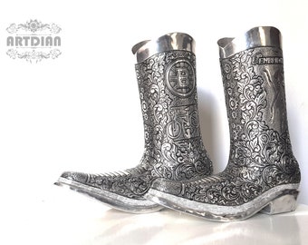 May include: A pair of silver-colored cowboy boots with intricate floral and geometric patterns. The boots feature a pointed toe, a decorative heel, and a cuff at the top. The boots have a metallic sheen and are set against a white background.