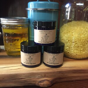 May include: Three glass jars with lids containing various ingredients, including a jar of yellow wax beads, a jar of clear liquid, and a jar of sugar scrub. The sugar scrub jars are labeled "Melken Mutter Farm" and have a drawing of a pig on them. The jars are sitting on a wooden surface.
