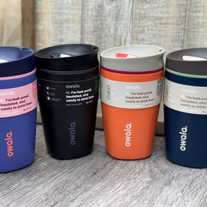 May include: Four reusable coffee cups in different colors: blue, black, orange, and navy blue. Each cup has a white label with the text "owala" and the phrase "Hi, I'm leak proof, insulated, and comfy to drink from."