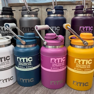 May include: A collection of RTIC Half Gallon Outback Jugs in various colors: black, gray, navy, purple, white, teal, pink, and yellow. Each jug has a handle and a screw-on lid. The jugs are designed for better tasting beverages.