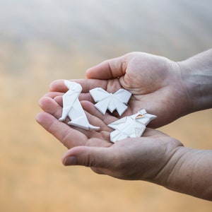 Pu&ograve; includere: Tre figure di carta origami bianca, una farfalla, un cane e una forma geometrica, sono tenute nei palmi di due mani.