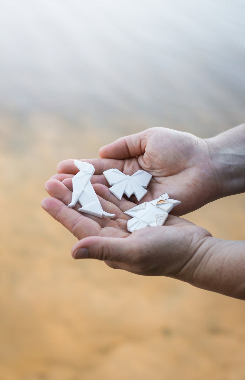 Pu&ograve; includere: Tre figure di carta origami bianca, una farfalla, un cane e una forma geometrica, sono tenute nei palmi di due mani.