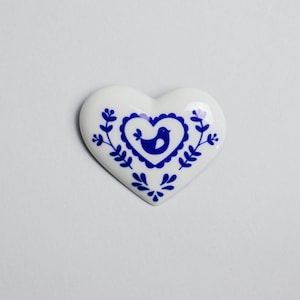 May include: White ceramic heart-shaped ornament with a blue bird design inside a heart. The bird has a blue body and white wings. The heart is surrounded by blue floral designs.