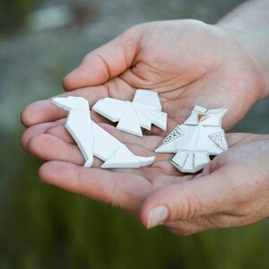 Pu&ograve; includere: Tre figure di origami in ceramica bianca, un cane, una farfalla e un gufo, sono tenute nel palmo della mano di una persona. Il gufo ha accenti dorati.