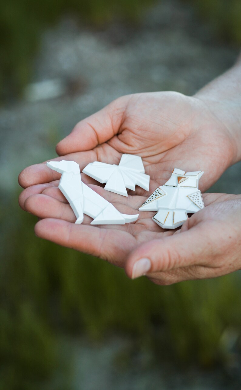 Pu&ograve; includere: Tre figure di origami in ceramica bianca, un cane, una farfalla e un gufo, sono tenute nel palmo della mano di una persona. Il gufo ha accenti dorati.