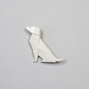 Pu&ograve; includere: Scultura di cane origami in ceramica bianca. Il cane &egrave; seduto con la testa inclinata verso l'alto e la coda arrotolata dietro di lui.