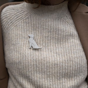 Pu&ograve; includere: Una spilla a forma di cane origami bianco appuntata su un maglione beige.