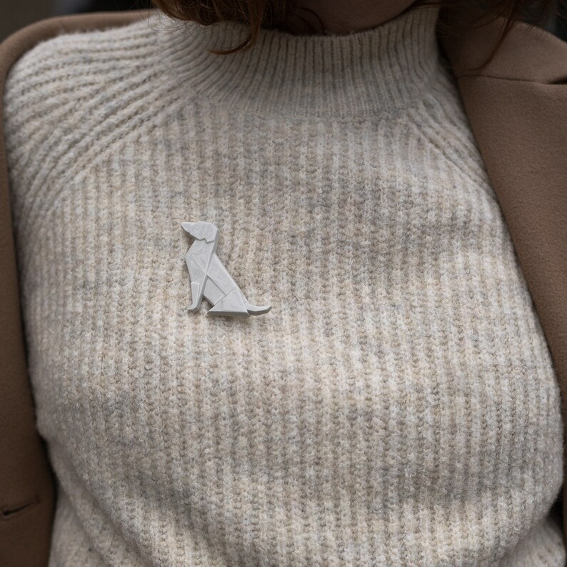 Pu&ograve; includere: Una spilla a forma di cane origami bianco appuntata su un maglione beige.