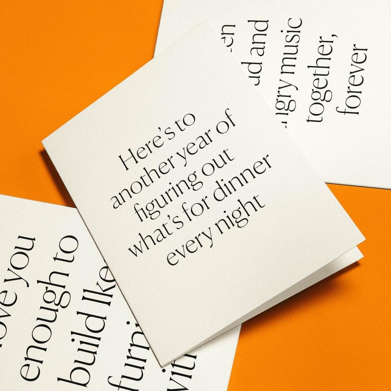 May include: White greeting cards with black text on an orange background. The cards have phrases like "Here's to another year of figuring out what's for dinner every night." Other cards have phrases like "love you enough to build like" and "hungry music together, forever."