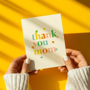 K&ouml;nnte beinhalten: Eine cremefarbene Gru&szlig;karte mit den Worten "thank you mom" in bunten, verspielten Buchstaben. Die Karte wird von H&auml;nden gehalten, die einen wei&szlig;en Pullover vor einem leuchtend gelben Hintergrund tragen.