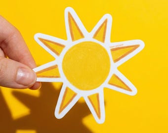 May include: A yellow sun sticker with a white border is held against a bright yellow background. The sun has a round center and eight pointed rays. The text "paper and stuff" is printed on the sticker.