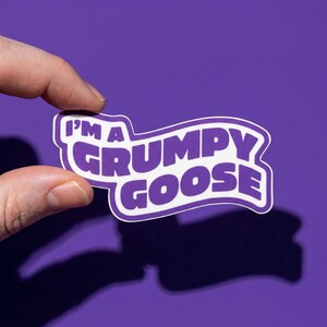 May include: A purple sticker with the text "I'M A GRUMPY GOOSE" in white, outlined in purple. The sticker is held between fingers against a purple background. The sticker's design is a playful, cartoonish style.