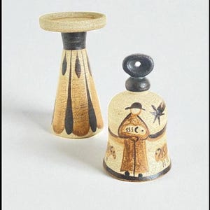 May include: Two ceramic decorative items. One is a candlestick with a beige top and a black and beige patterned base. The other is a bell-shaped item with a black top and a beige body with a painted figure and star.
