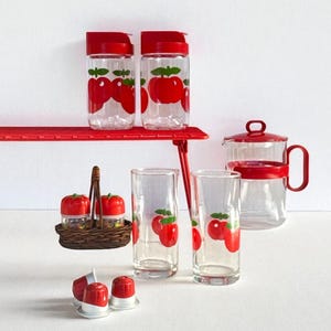 Könnte beinhalten: Eine Sammlung von Küchenutensilien mit einem roten Apfel-Thema. Enthält klare Gläser, eine Teekanne, Gewürzgläser und kleine Behälter, alle mit roten Apfelmotiven verziert. Die Artikel sind auf einem roten Regal und einer weißen Oberfläche angeordnet.