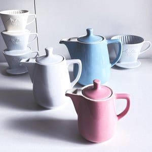 Puede incluir: Una colección de teteras y vertedores de café de cerámica en varios colores pastel. Las teteras son blancas, celeste y rosas. Los vertedores son blancos y celeste. Los artículos están colocados sobre una superficie blanca.