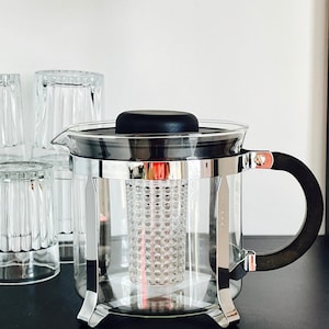 Może przedstawiać: Szklany czajnik do herbaty z czarnym uchwytem i pokrywką ze stali nierdzewnej. Czajnik ma wbudowany filtr i stoi na czarnej powierzchni. Obok czajnika stoi cztery przezroczyste szklanki ułożone w stos.