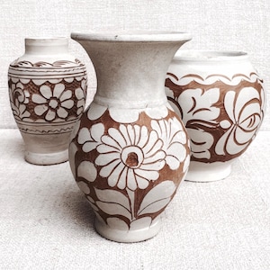 May include: Three ceramic vases with a white and brown floral design. The vases vary in shape and size, with the central vase being the largest. The floral patterns are hand-painted, adding a unique touch to each piece. The vases are displayed on a white surface.
