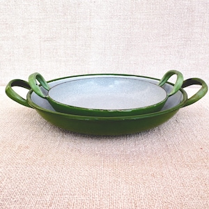 May include: Two green enamelware pans, one nested inside the other. The pans have a rounded shape with a wide rim and two curved handles. The interior of the pans is a light gray color.
