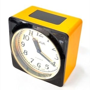 May include: A vintage square alarm clock in a black and yellow case. The clock face is white with black numerals and hands. The clock reads "LAVA" and "11 Jewels" and "Made in USSR".