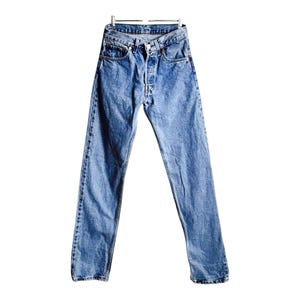 Könnte beinhalten: Hellblaue Jeans aus Denim, die an einem Metallbügel hängen. Die Jeans haben einen Knopfverschluss, ein klassisches Fünf-Taschen-Design und eine verwaschene Waschung. Der Straight-Leg-Stil ist sichtbar. Die Jeans sind vor weißem Hintergrund.