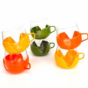 Op de afbeelding: Set van zes glazen bekers met oranje en groene kunststof houders. De bekers zijn in paren gestapeld, met de houders die van kleur wisselen.