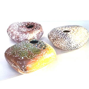 May include: Three ceramic vases in different colors and patterns. The vases are shaped like rounded stones with a hole in the top for flowers. The vases are white with brown, yellow, and green speckles.