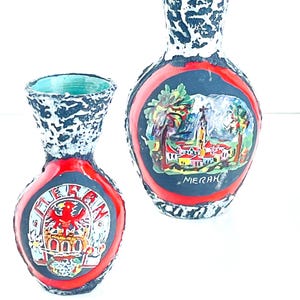 May include: Two ceramic vases with a black and white speckled glaze. The larger vase has a red rim and a painted scene of a town with the word "MERAN" below. The smaller vase has a red rim and a painted crest with a red eagle and the word "DEUTSCHLAND" around it.