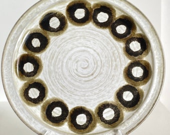 Op de afbeelding: Een ronde keramische plaat met een crèmekleurige achtergrond en een spiraalvormig ontwerp in het midden. De plaat heeft een ring van donkerbruine cirkels met witte centra aan de rand. De plaat wordt gepresenteerd op een transparante plastic standaard.
