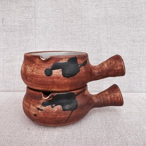 Puede incluir: Dos cuencos de cerámica marrón con salpicaduras de pintura negra y un asa a cada lado. Los cuencos están apilados uno encima del otro.