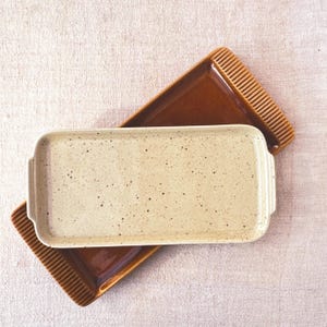 May include: Two rectangular ceramic serving trays. The top tray is beige with brown speckles and has rounded edges. The bottom tray is brown with a textured edge.