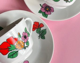 May include: Two white teacups with floral designs. The cups have red, orange, yellow, pink, and green flowers. The cups are on white saucers with matching floral designs.