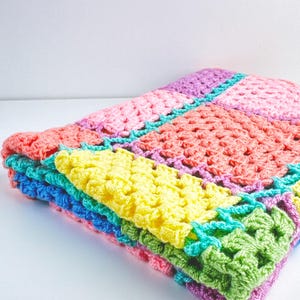 Könnte beinhalten: Eine bunte Häkeldecke mit Quadraten in den Farben Rosa, Gelb, Grün, Blau und Lila. Die Decke ist zu einem Drittel gefaltet und die Kanten sind sichtbar.