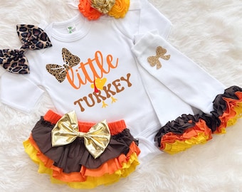 May include: A white baby bodysuit with the words "little TURKEY" in orange and brown. The outfit includes a matching headband with orange and gold flowers, a pair of leopard print bows, ruffled bloomers, and white socks with gold bows. The outfit is designed for a baby.