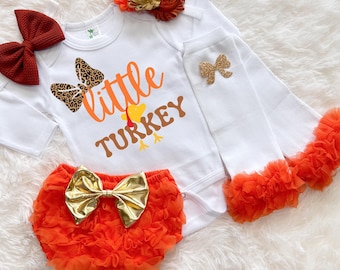 May include: A white baby bodysuit with the text "little TURKEY" in orange and brown, with a turkey graphic. The set includes orange ruffled bloomers with a gold bow, white leg warmers with orange ruffles, and a burgundy bow. A floral headband completes the outfit.