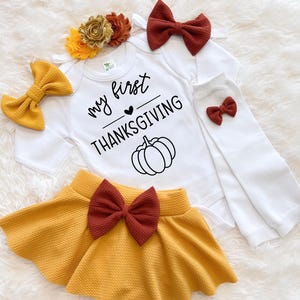 Könnte beinhalten: Ein Baby-Thanksgiving-Outfit-Set mit einem weißen Body mit dem Text "my first THANKSGIVING" und einer Kürbisgrafik. Enthält einen senfgelben Rock, Schleifen und ein Blumenstirnband. Das Set ist für das erste Thanksgiving eines Babys gedacht.