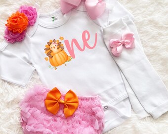 May include: A white baby bodysuit with the word "one" in pink script and a pumpkin-themed graphic. The set includes a pink ruffled diaper cover with an orange bow, a large pink bow, and white leg warmers with a small pink bow. A floral headband is also included.