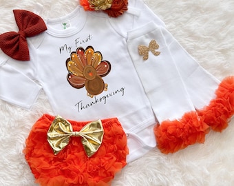 May include: A white baby bodysuit with a turkey graphic and the text "My First Thanksgiving." The set includes orange ruffled bloomers with a gold bow, white leg warmers with orange ruffles, a burgundy bow, and a floral headband.