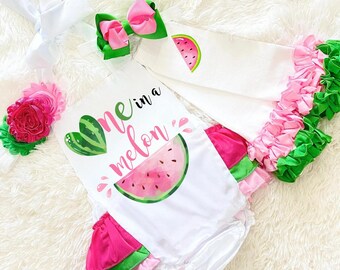 以下が含まれることがあります： スイカをテーマにした白いベビー服セット。セットには、「one in a melon」の文字が入ったロンパース、ピンクの花のヘアバンド、フリル付きブルマが含まれています。衣装はピンクとグリーンのアクセントがあります。