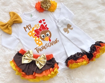 May include: A white baby bodysuit with the text "My 1st Thanksgiving" and a turkey graphic. The set includes a ruffled diaper cover, bow, headband, and arm warmers in gold, brown, orange, and yellow. The outfit is designed for a baby.