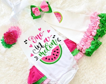 以下が含まれることがあります： 「One in a melon」の文字が入った、スイカをテーマにした白いベビー服。 ロンパース、レッグウォーマー、ヘアバンドが含まれています。 ロンパースにはスイカのスライスグラフィックがあります。 レッグウォーマーとリボンは、ピンクとグリーンのフリルでトリミングされています。