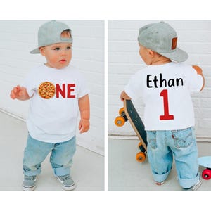 May include: A white t-shirt with a pizza graphic and the word "ONE" in red. The back of the shirt has the name "Ethan" and the number "1" in red. The child is wearing blue jeans and a gray baseball cap.