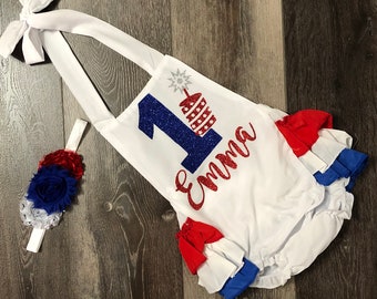 May include: White onesie with red, white, and blue ruffles. The onesie has a glittery blue number one with a red, white, and blue firework graphic. The onesie says "Emma" in red glitter. A matching headband with red, white, and blue fabric flowers is included.