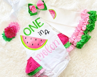 以下が含まれることがあります： ウォーターメロンのグラフィックと「ONE in a melon」のテキストが書かれた白いロンパース。ロンパースの裾にはピンクとグリーンのフリルが付いています。ピンクとグリーンのリボンと、ピンクの花が付いたヘッドバンドも含まれています。